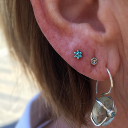 A small blue Anatometal flower is worn in a lobe piercing, the third one up. the lower two have a silver stud and a ring with a hanging stone