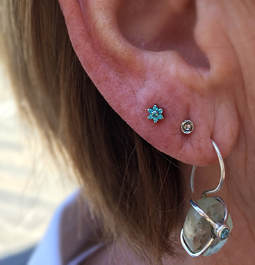 A small blue Anatometal flower is worn in a lobe piercing, the third one up. the lower two have a silver stud and a ring with a hanging stone