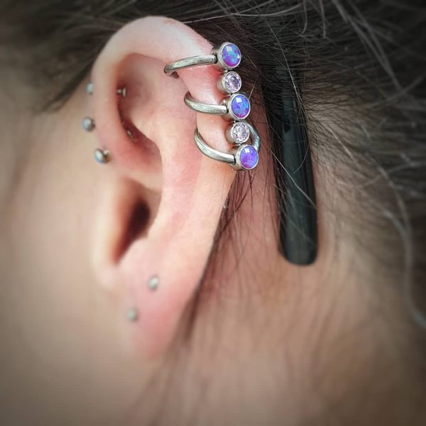 three forward helix piercings with purple opals and a further three helix piercings wearing custom Anatometal jewellery. The piece is five purple gems joined together with three rings wrapping through the piercings