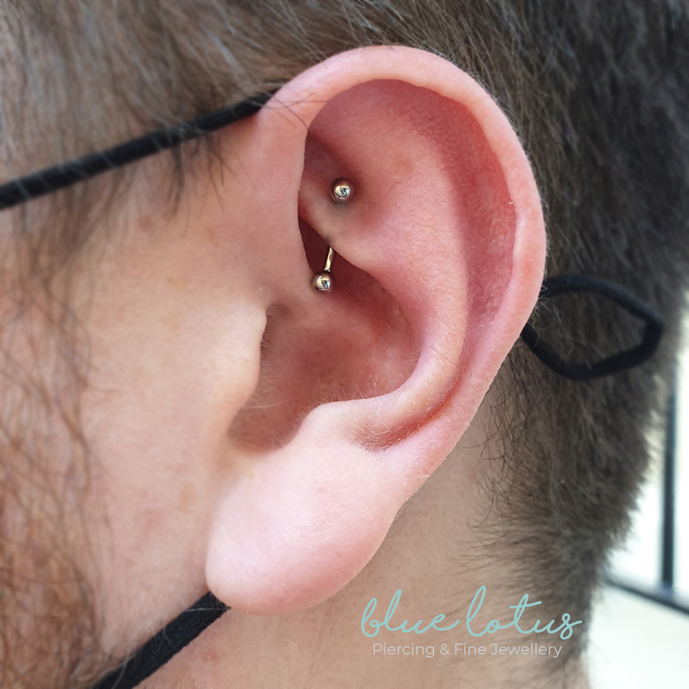 A high polished curved bar in a new rook piercing, there is a black mask line wrapping the ear and the studios logo to the bottom