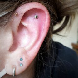 clients own silver ring in lobe, two stacked (one above the other) lobe piercings wear aquamarine crystals. there is a helix and forwrad helix wearing a Hera and Tribead attachment from Anatometal