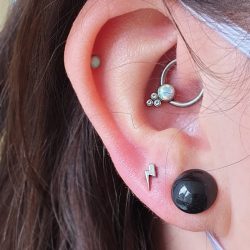 a hammered lightning bolt shape from Anatometal in a lobe piercing, as well as an opal captive cluster in a daith piercing ring. there is also a small white opal poking out from a helix piercing and a black stretcher in the lower lobe