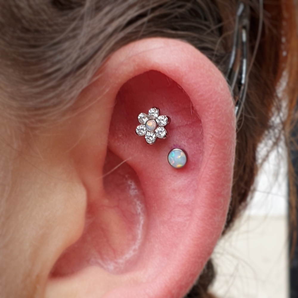 An Anatometal flower with clear gem petals and an opal cabochon centre on teh flat of an ear, right beside a flat opal cabochon